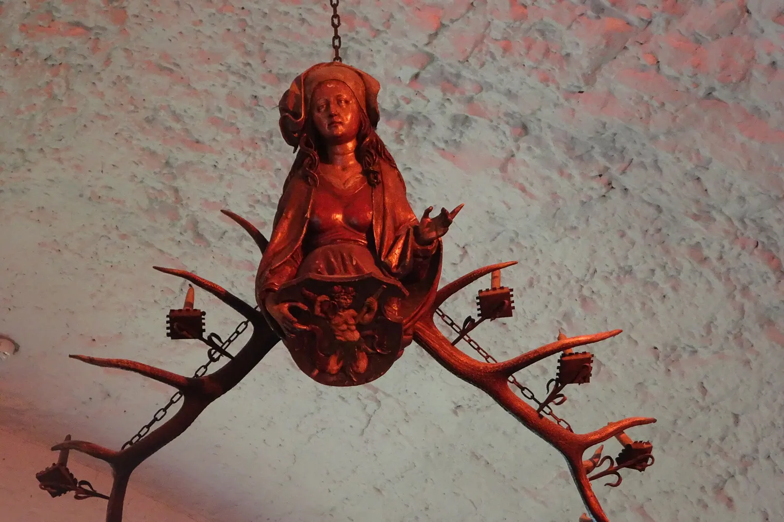 A carved wooden figure of a woman set between two branching antlers, forming a chandelier with candle holders, hanging from a whitewashed ceiling