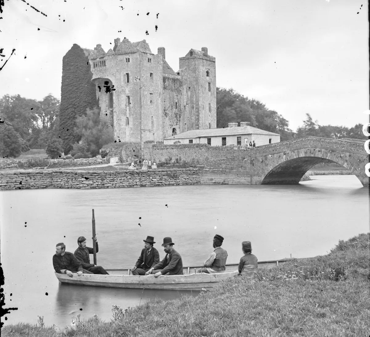 A black-and-white photograph of Bunratty Castle behind a stone bridge, with a rowing boat and men in dark coats in the foreground
