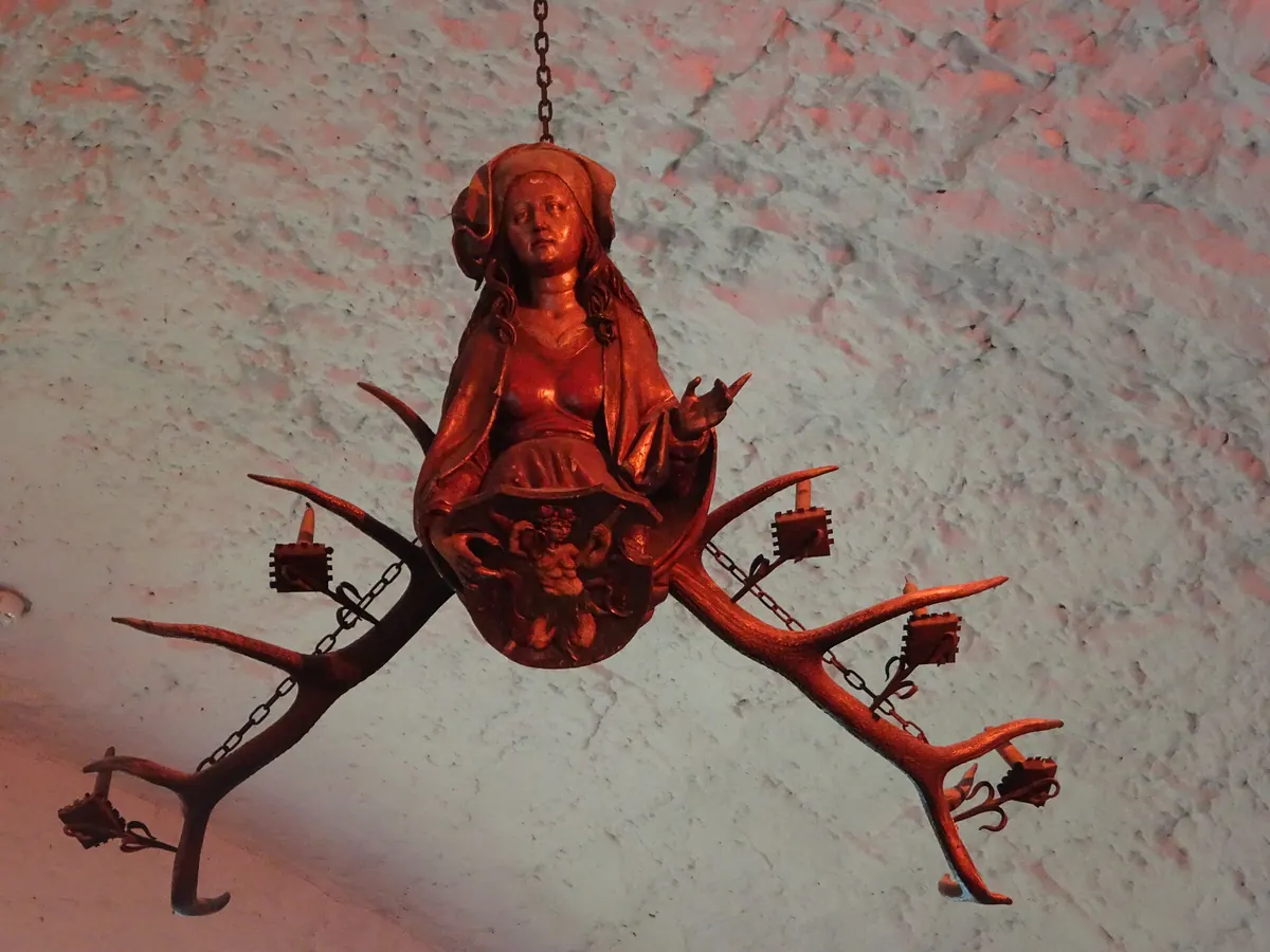 A carved wooden figure of a woman between two antlers, forming a candle chandelier hanging from a whitewashed ceiling