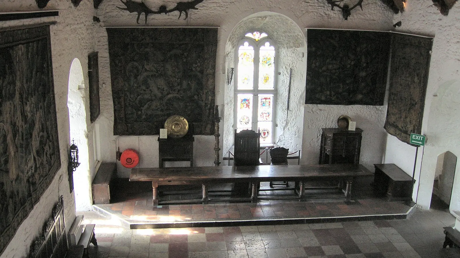 The Great Hall of Bunratty Castle in daylight, a long wooden table on a raised dais beneath large tapestries and a stained-glass window, with antlers mounted high on the whitewashed walls