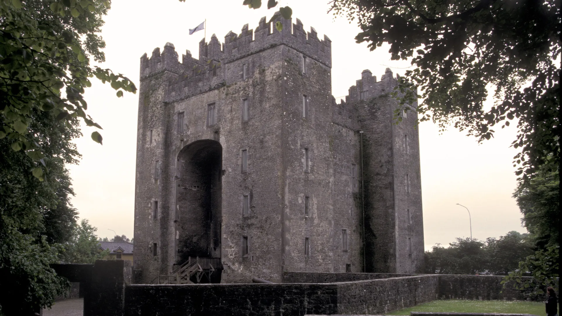 Bunratty Castle at dusk, a grey stone tower house with battlements and a tall arched recess, framed by dark trees, with a flag flying above the roof